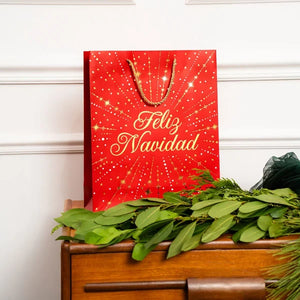 Bolsa de regalo roja Feliz Navidad con letras y destellos dorados, colocada sobre un mueble de madera junto a ramas verdes navideñas.
